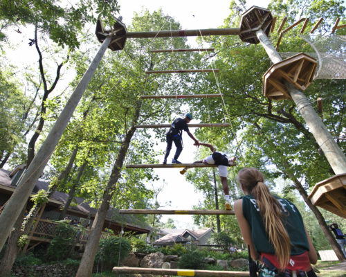 A friend giving another a helping hand to ascend a large ropes obstacle course
